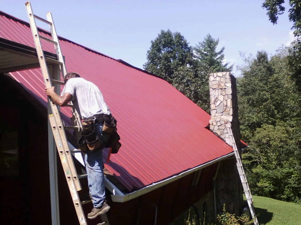 Roofer on a ladder beside a red metal roof and stone chimney — Soffit & Fascia Repair by Roofing Brothers in Colo